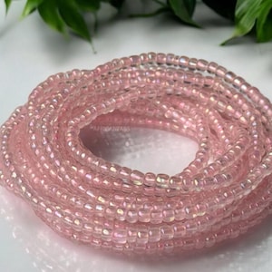 May include: A close-up of a pink beaded waist beads, also known as waist beads or belly beads. The beads are iridescent and have a subtle shimmer. The beads are wrapped around each other in a spiral shape.