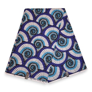 Op de afbeelding: Een gevouwen stuk stof met een herhalend patroon van blauwe, lichtblauwe en witte spiraalvormige schelpvormen. Het textiel heeft een bedrukt ontwerp met de tekst "VERITABLE BLOCK WAX PRINT" zichtbaar langs de onderrand van de stofvouw.