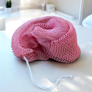 Könnte beinhalten: Ein rosa Duschschwamm aus Netzstoff mit einer weißen Schnurschlaufe. Der Schwamm hat eine leuchtend rosa Farbe und ein strukturiertes, netzartiges Aussehen. Er liegt auf einer weißen Oberfläche, was auf eine Badezimmerumgebung hindeutet.