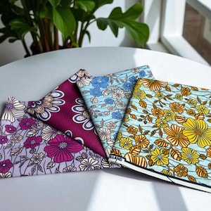 Op de afbeelding: Vier gevouwen stoffen vierkanten met bloempatronen. De stoffen hebben verschillende kleuren, waaronder paars, blauw en geel, met witte en bruine accenten. De bloemmotieven zijn gedetailleerd en bedekken het hele oppervlak van de stof.
