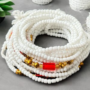May include: White beaded waist beads with red and gold accents. The beads are strung on a white cord and are arranged in a circular shape.