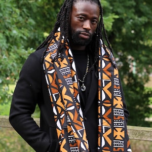Op de afbeelding: Een man met dreadlocks draagt een zwart blazer met een kleurrijke, gepatroneerde sjaal. De sjaal heeft een geometrisch ontwerp in zwart, wit, oranje en bruin.