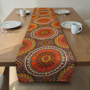 Op de afbeelding: Een decoratieve tafelloper met een herhalend cirkelvormig patroon in tinten oranje, geel en bruin. Het ontwerp heeft ingewikkelde geometrische vormen en een gestippelde rand. De loper ligt op een houten tafel, met witte borden en mokken.