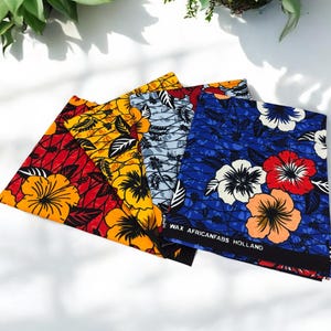 Op de afbeelding: Drie stofstalen met bloemenpatronen. De stalen zijn rood, blauw en grijs met gele bloemen. De stof is geëtiketteerd met "WAX AFRICANFABS HOLLAND".