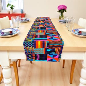 Op de afbeelding: Een kleurrijke tafelloper met een patchwork ontwerp. De loper heeft verschillende geometrische patronen in blauw, paars, oranje, roze en geel. Hij ligt op een lichtgekleurde houten tafel, gedekt voor een maaltijd.