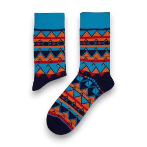 Op de afbeelding: Een paar sokken met patroon met een blauwe boord en teen. De sokken hebben een herhalend geometrisch ontwerp in oranje, rood en blauw op een marineblauwe achtergrond. De sokken worden op een witte achtergrond gepresenteerd.