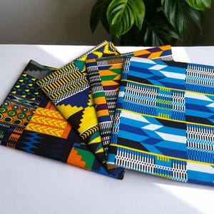 Op de afbeelding: Een verzameling kleurrijke Afrikaanse waxprint stoffen. De stoffen hebben geometrische patronen in blauw, geel, oranje en groen. De stoffen zijn gevouwen en op een wit oppervlak geplaatst.