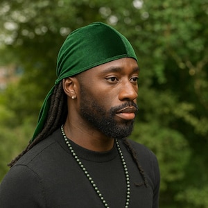 May include: A man wearing a dark green velvet durag with long dreadlocks and a black shirt with a dark green beaded necklace.