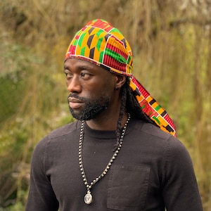 Puede incluir: Un hombre con una camisa negra, un collar de cuentas plateadas y una bandana de colores con un patrón. La bandana está anudada en la parte posterior del cuello y los extremos cuelgan por la espalda.