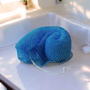 Op de afbeelding: Een opgerolde, helderblauwe mesh-badspons met een wit touwtje eraan. De spons ligt op een wit oppervlak, met een witte badkuip op de achtergrond.