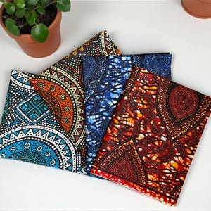 Op de afbeelding: Vier gevouwen stoffen stukken met levendige Afrikaanse wasprintpatronen. De stoffen hebben geometrische en bloemmotieven in tinten blauw, oranje, rood en wit. Een potplant is zichtbaar op de achtergrond.