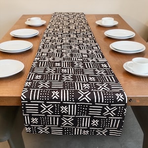 Puede incluir: Un camino de mesa con estampado en blanco y negro con diseños geométricos se muestra sobre una mesa de madera. Platos y tazas blancos están dispuestos alrededor del camino de mesa, lo que sugiere un entorno de comedor. El patrón del camino de mesa añade un toque decorativo.