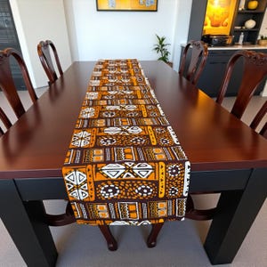 Op de afbeelding: Een rechthoekige tafelloper met een herhalend geometrisch patroon in bruin, oranje en wit. De loper ligt op een donkerbruine houten tafel. De tafel wordt omringd door donkerbruine houten stoelen.
