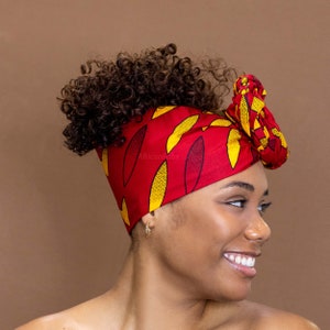 Puede incluir: Un turbante rojo y amarillo con estampado y un gran lazo en la parte superior. El turbante está atado alrededor de la cabeza de una persona y el lazo está colocado en el lado derecho de la cabeza.