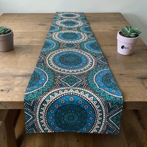 Op de afbeelding: Een rechthoekige tafelloper met een herhalend cirkelvormig patroon in tinten teal, turkoois en wit. De loper ligt op een houten tafel, met vetplanten in potten aan weerszijden.