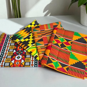 Può includere: Una collezione di tessuti africani stampati a cera colorati. I tessuti presentano motivi geometrici in rosso, giallo, nero e verde. I tessuti piegati sono disposti su una superficie bianca, mostrando i disegni vivaci.