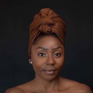 Peut inclure: Une femme à la peau brune foncée porte un turban marron. Elle regarde directement la caméra avec une expression neutre. Elle porte un collier en or et de petites boucles d'oreilles.