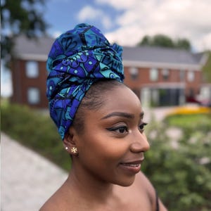 Op de afbeelding: Een vrouw draagt een blauw en turquoise patroon hoofddoek. De hoofddoek heeft een gedetailleerd ontwerp met bloemen en geometrische vormen. De vrouw draagt gouden oorstekers en heeft donkere eyeliner. Op de achtergrond zijn een gebouw en groen te zien.