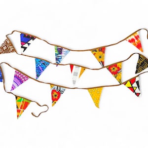 Puede incluir: Una guirnalda de tela colorida con banderas triangulares en varios patrones y colores, incluyendo rojo, amarillo, azul, verde y negro. La guirnalda está enhebrada en un cordón marrón y está colgando sobre un fondo blanco.