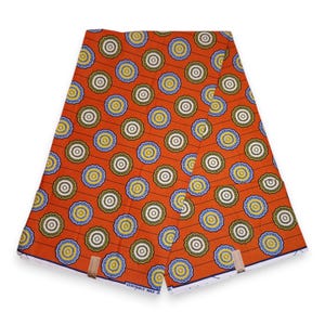 May include: Orange fabric featuring a repeating pattern of circular designs in white, yellow, blue, and green. The fabric is folded and held together with wooden clips. The text "VERITABLE WAX" is visible along the edge.