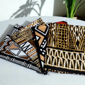 Op de afbeelding: Een verzameling gevouwen stoffen met gedurfde geometrische patronen in zwart, wit, bruin en goud. De stoffen zijn in een waaier vorm gerangschikt, waarbij de verscheidenheid aan ontwerpen wordt getoond. De algehele esthetiek is levendig en cultureel geïnspireerd.