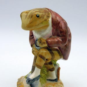 May include: A ceramic figurine of an anthropomorphic frog wearing a dark red jacket, a white cravat, and tan trousers with brown horizontal stripes. The frog is leaning forward while holding a shovel, standing on a textured, light-coloured base.