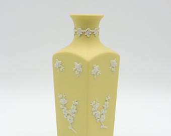 May include: A pale yellow, square vase with white floral relief designs. The vase has a decorative band around the neck and floral patterns on the body. The vase is a decorative item.