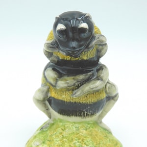 May include: A ceramic figurine of a bumblebee with a glossy black head, yellow and black striped body, and light grey limbs. The bee is depicted in a seated position with arms crossed, resting on a textured, light green, rounded base.