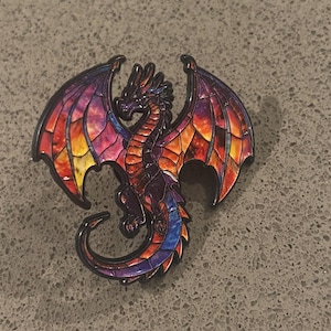 Könnte beinhalten: Eine Metall-Emaille-Anstecknadel in Form eines Drachen, dessen Flügel und Körper einen farbenfrohen Buntglaseffekt aufweisen. Der Drache hat eine dunkle Umrandung und zeigt leuchtende Violett-, Orange-, Rot- und Blautöne in den Flügeln.