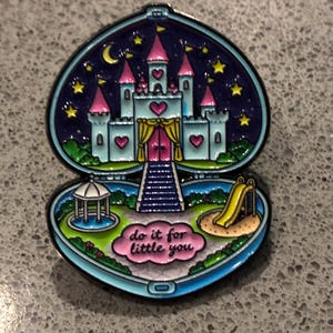May include: A heart-shaped enamel pin featuring a light blue castle with pink roofs and heart accents against a dark blue starry night sky. The bottom section shows a green park with a gazebo, a yellow slide, and a pink cloud with the text do it for little you.