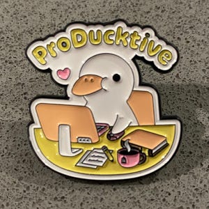 以下が含まれることがあります： 「ProDucktive」の文字が黄色で書かれた白いアヒルのピンバッジ。アヒルはパソコン、コーヒー、本のある机に座っています。ピンバッジは黄色の縁取りとピンクのハートが付いています。