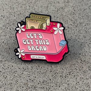 May include: A pink enamel pin in the shape of a toaster, featuring the text "LET'S GET THIS BREAD". The toaster has cash protruding from the slot. The pin is decorated with white flowers and stars. The words "HOT & FRESH" are also on the pin.