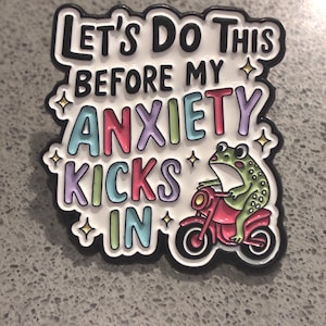 May include: A white enamel pin with black outlines featuring a green frog riding a red scooter. The text reads LET'S DO THIS BEFORE MY ANXIETY KICKS IN in colourful letters. Small yellow star accents are scattered around the text on the white background.