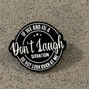 Peut inclure: Une broche émaillée noire circulaire avec du texte blanc qui indique IF WE ARE IN A Don't Laugh SITUATION DO NOT LOOK OVER AT ME, avec trois petites étoiles blanches centrées au-dessus du mot Don't dans une police cursive stylisée.