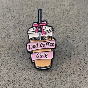 Op de afbeelding: Een metalen emaille speld in de vorm van een plastic beker ijskoffie met een rietje, een roze strik en een roze banner om het midden met de zwarte tekst Iced Coffee Girly in een schrijflettertype tegen een lichtbruine vloeibare achtergrond.