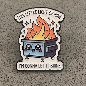 Peut inclure: Une broche en émail métallique représentant une benne à ordures bleu clair avec un visage mignon, remplie de flammes orange et jaune vif. Le texte "THIS LITTLE LIGHT OF MINE" est en haut et "I'M GONNA LET IT SHINE" est en bas en noir.