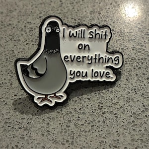 Peut inclure: Une broche en émail métallique représentant un pigeon de dessin animé avec un corps gris et une tête noire. Le pigeon a de grands yeux écarquillés et se tient à côté d'une bulle blanche avec le texte noir, I will shit on everything you love.