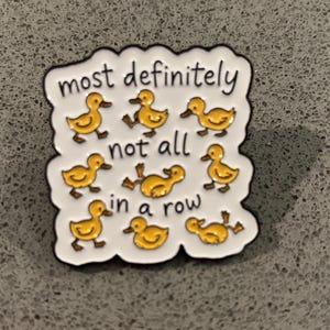 May include: A white enamel pin with a black border featuring eight yellow ducklings in various poses and the black text that reads most definitely not all in a row. The pin has a wavy, cloud-like edge and a glossy finish on the surface.