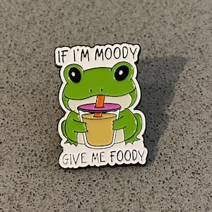 May include: A green frog enamel pin holding a drink with a straw. The pin has the text "IF I'M MOODY GIVE ME FOODY" in black letters. The frog has large black eyes and is outlined in black.