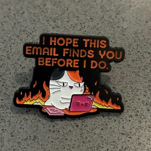 Könnte beinhalten: Eine schwarze Emaille-Anstecknadel mit einer Glückskatze, die an einem rosa Laptop arbeitet, umgeben von orangefarbenen und gelben Flammen. Der Text über der Katze lautet, I HOPE THIS EMAIL FINDS YOU BEFORE I DO. Schwarzer Hintergrund, orange Schrift.
