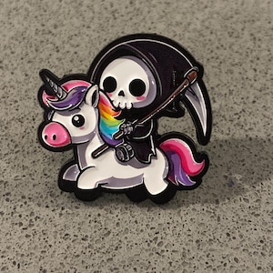 Peut inclure: Une broche en émail métallique représentant une licorne blanche avec une crinière et une queue arc-en-ciel, portant une petite faucheuse vêtue d'une cape noire à capuche. La faucheuse tient une faux argentée au manche marron, museau rose.