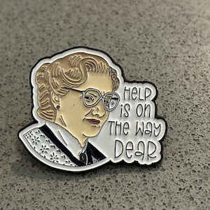 Peut inclure: Une broche en émail métallique représentant une illustration stylisée d'une personne âgée aux cheveux brun clair, portant de grandes lunettes et un col blanc à motifs. Le texte "HELP is on THE WAY DEAR" est inscrit en noir sur un fond blanc.