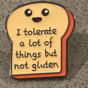 May include: A novelty enamel pin in the shape of a slice of bread, featuring a smiling face and the words "I tolerate a lot of things but not gluten." The pin has a yellow centre, an orange border, and a silver outline.