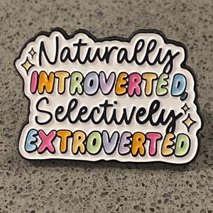 May include: A colourful enamel pin with the words "Naturally Introverted Selectively Extroverted" in a playful font. The pin has a black outline and features a variety of colours, including pink, orange, blue, and yellow.