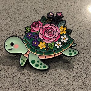 May include: A green turtle-shaped enamel pin with a floral design. The turtle has a smiling face and is decorated with pink, purple, and yellow flowers. The pin has a black outline.