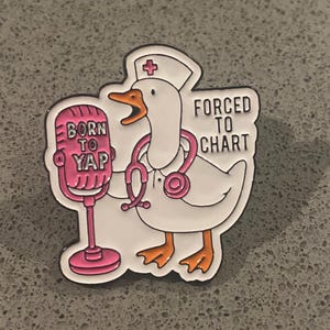 May include: A white enamel pin featuring a goose dressed as a nurse with a pink stethoscope and cap. The goose stands next to a pink microphone with the text BORN TO YAP. The text FORCED TO CHART is printed in black on the right side of the pin.
