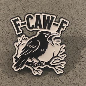May include: A black and white enamel pin featuring a crow perched on a branch with its beak open. Above the bird, the text F-CAW-F is displayed in a bold, stylised font. The design uses high-contrast black lines against a white background.
