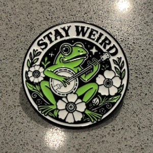 May include: A circular enamel pin featuring a green frog playing a banjo, surrounded by white flowers and black foliage. The text STAY WEIRD is printed in black capital letters along the top edge of the pin against a white background border.