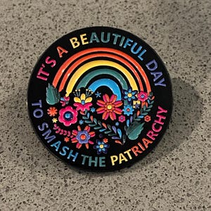 May include: Black circular pin with a rainbow and floral design. The pin features the text "It's a beautiful day to smash the patriarchy" in a colorful font. The pin has a glossy finish.