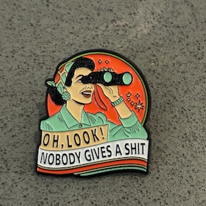 Peut inclure: Un pin's en émail circulaire représentant une personne aux cheveux noirs portant un foulard vert, regardant à travers des jumelles noires. Le fond est orange avec des étoiles. Une bannière blanche affiche le texte : OH, LOOK! NOBODY GIVES A SHIT.
